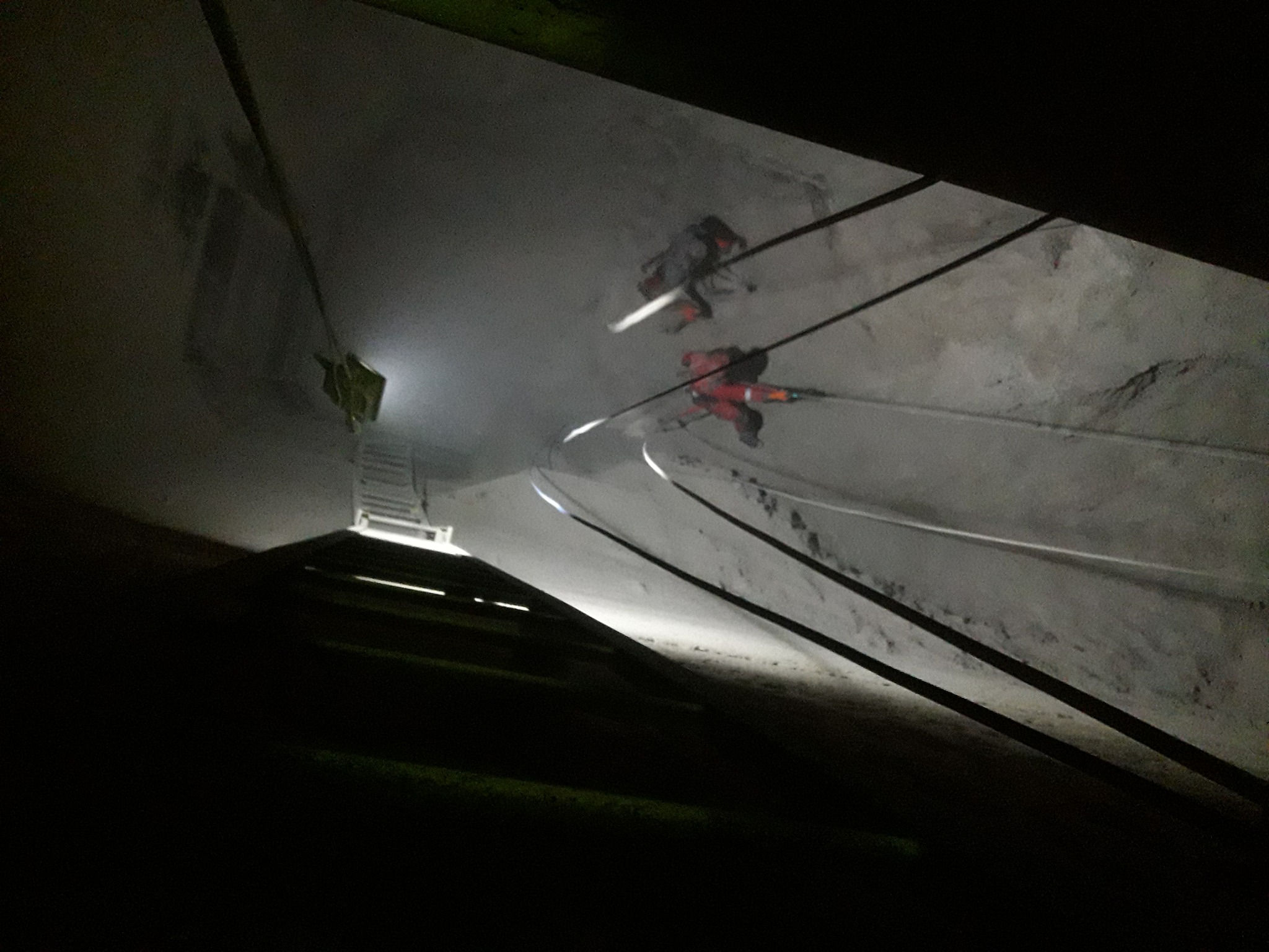 Dwóch pracowników w kombinezonach, zabezpieczonych linami, wykonuje prace na wysokościach na szarej, chropowatej powierzchni, oświetlonych punktowo. W tle widoczna drabina.