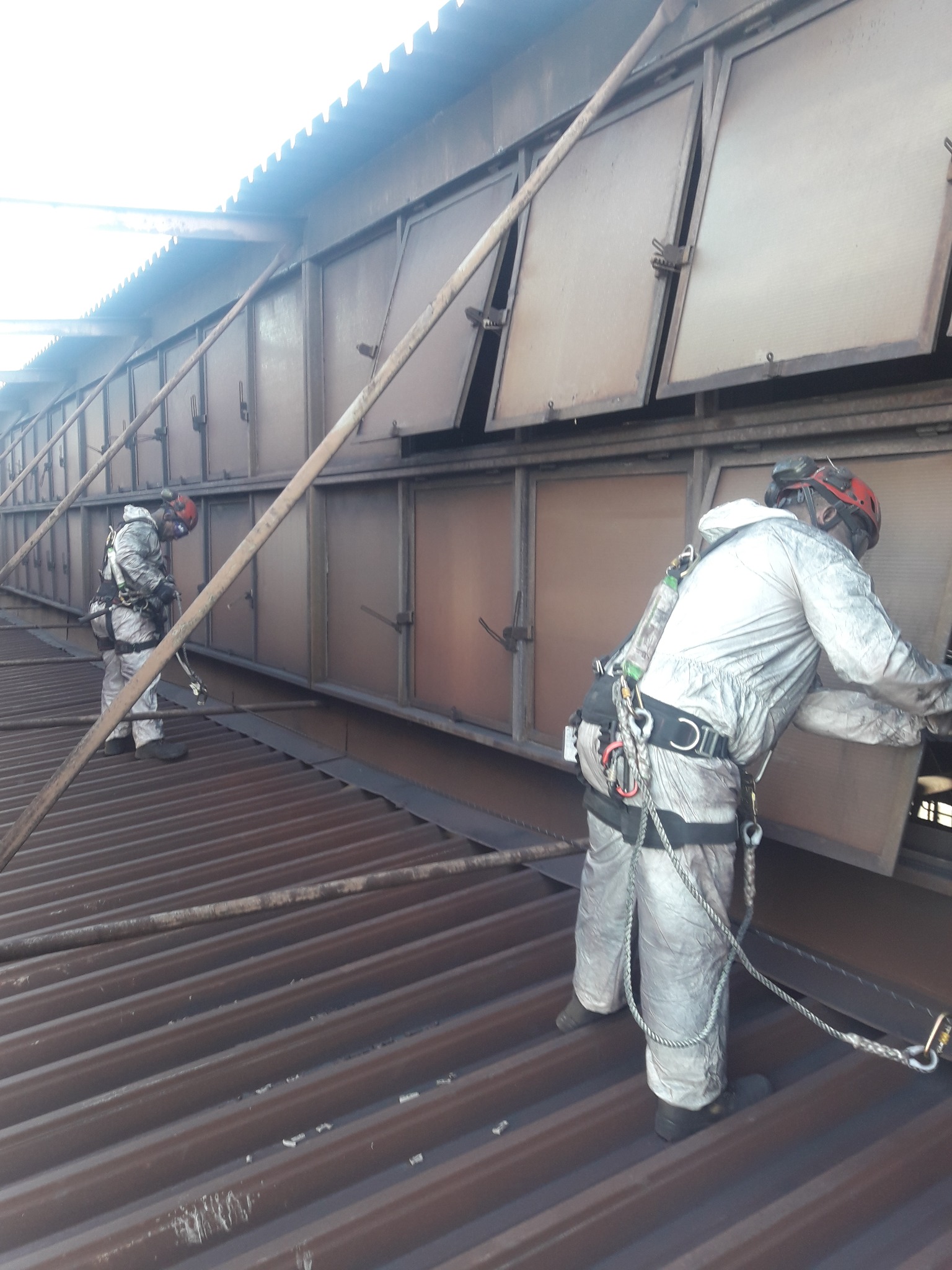 Dwóch pracowników w kombinezonach ochronnych i kaskach, zabezpieczonych linami, wykonuje prace na wysokości na dachu budynku przemysłowego, przy otwartych metalowych klapach wentylacyjnych.