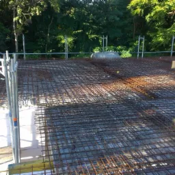 Zbrojenie stropu na budowie domu jednorodzinnego. Widoczne białe bloczki, rusztowanie i pracownik w oddali. Perspektywa z g&oacute;ry ukazuje siatkę zbrojeniową.