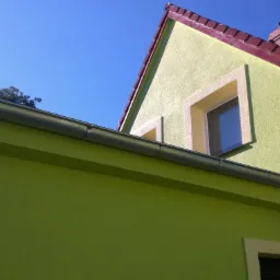 Fragment domu z zieloną elewacją, szarymi rynnami i czerwonym dachem, uchwycony pod kątem z błękitnym niebem w tle. Widoczne okno z jasną ramą.