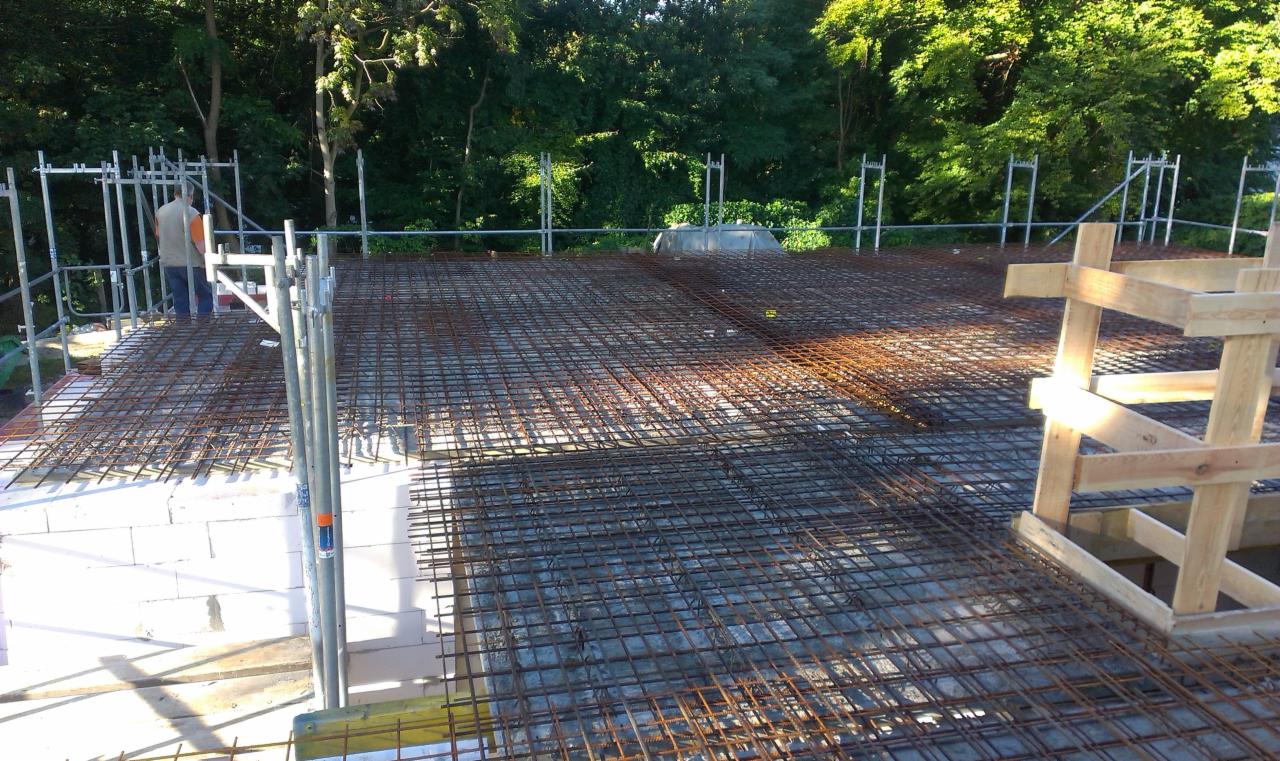Zbrojenie stropu na budowie domu jednorodzinnego. Widoczne białe bloczki, rusztowanie i pracownik w oddali. Perspektywa z góry ukazuje siatkę zbrojeniową.