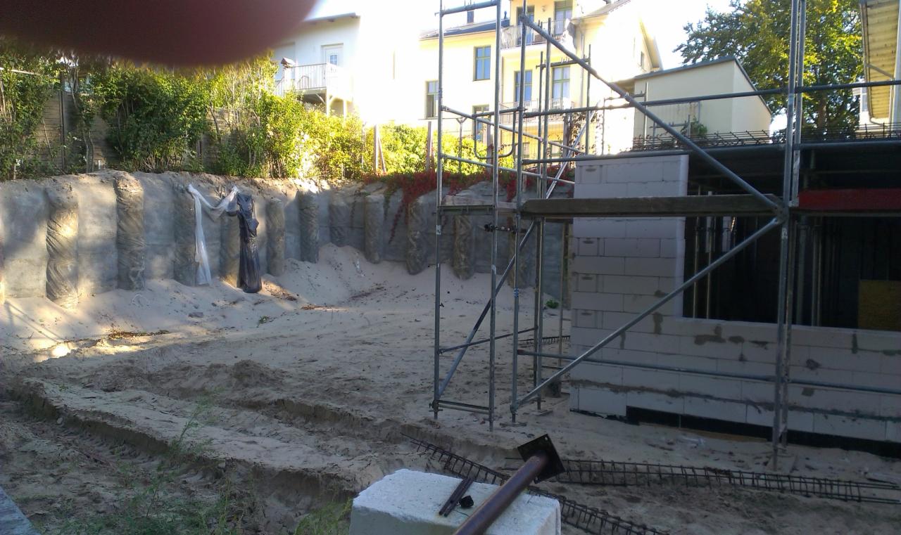 Budowa domu: fundamenty z bloczków, zabezpieczona skarpa, rusztowania. Widoczne zbrojenia i teren przygotowany pod dalsze prace. Dzień słoneczny.