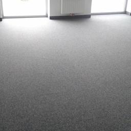 POLTARKIET Marcin Kuchnowski - Szara wykładzina dywanowa w jasnym pomieszczeniu z oknami i grzejnikiem. Ciemne listwy przypodłogowe. Widok na całą podłogę z perspektywy narożnika.