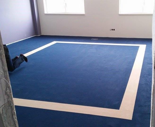 Niebieska wykładzina w pokoju z białym obramowaniem, widoczne nogi osoby pracującej przy montażu. Dwa okna w tle. Wykładzina świeżo po montażu.