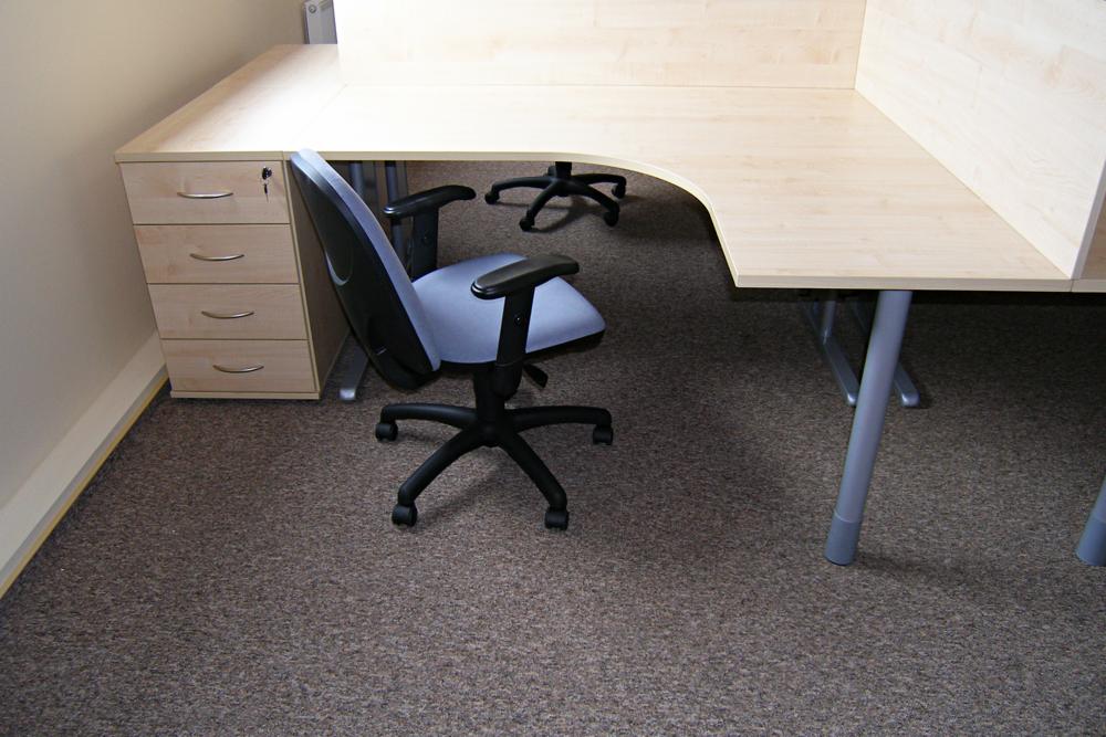 Wykładzina dywanowa w biurze pod blatem biurka z szufladami i krzesłem obrotowym. Widoczne fragmenty nóg biurka i innych krzeseł w tle.