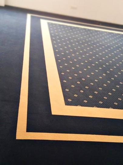 Granatowa wykładzina dywanowa z geometrycznym wzorem w beżowe kwadraty, obramowana beżową bordiurą. Widok z góry, fragment podłogi.