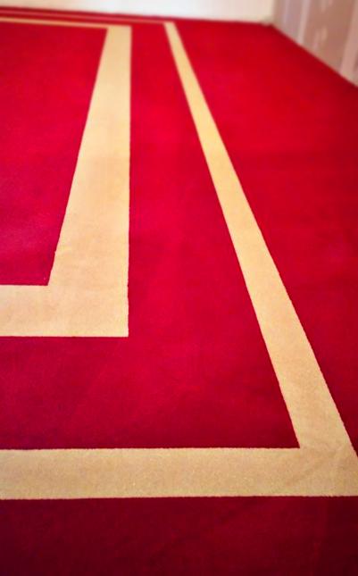 Czerwona wykładzina dywanowa z geometrycznym wzorem w odcieniach beżu, ułożona na podłodze. Widok z góry, bliski kadr.