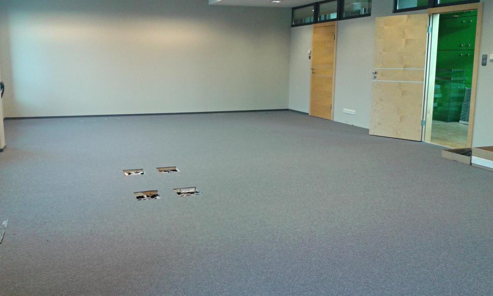 Szara wykładzina dywanowa w biurze z widocznymi otworami na podłodze, jasne ściany i drewniane drzwi w tle.