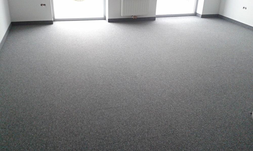 Szara wykładzina dywanowa w jasnym pomieszczeniu z oknami i grzejnikiem. Ciemne listwy przypodłogowe. Widok na całą podłogę z perspektywy narożnika.