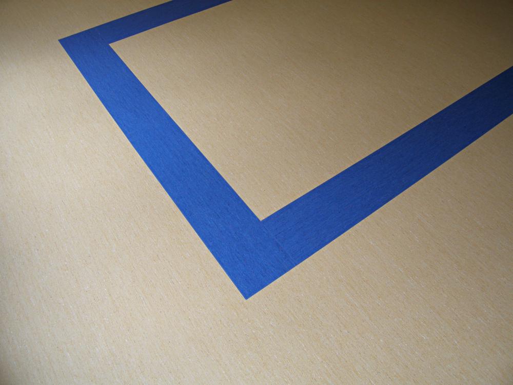 Jasna wykładzina z wyznaczonym niebieską taśmą malarską narożnikiem, przygotowana do montażu w pomieszczeniu. Widok z góry, delikatne oświetlenie.