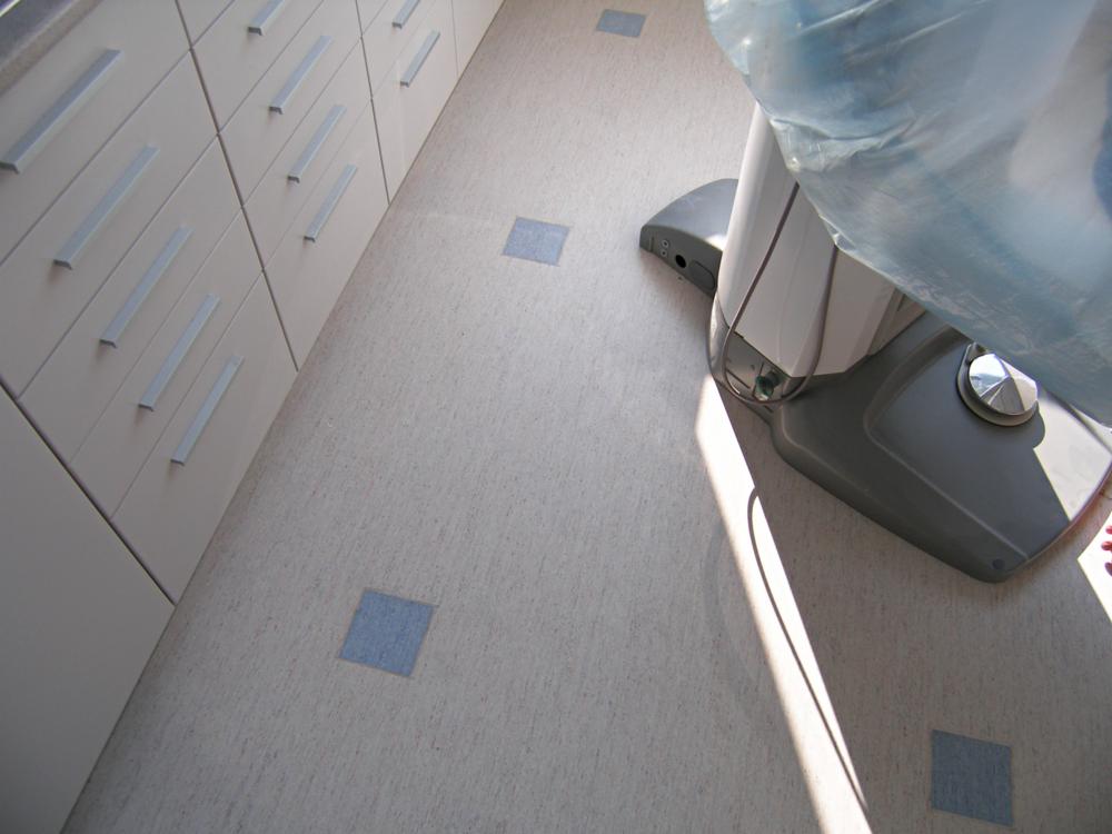 Jasna wykładzina z niebieskimi kwadratami w gabinecie, obok szafki i krzesło dentystyczne pod folią. Ukończona podłoga w pomieszczeniu usługowym.