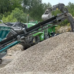 Kopalnia odkrywkowa: przesiewacz mobilny Powerscreen Warrior 800 i koparka ładują kruszywo na hałdę, w tle zielone drzewa.