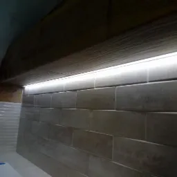 Ściana wyłożona beżowymi, prostokątnymi płytkami ceramicznymi imitującymi cegłę, oświetlona listwą LED ukrytą pod drewnianą belką sufitową.