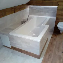 Nowoczesna wanna obudowana płytkami imitującymi cegłę z drewnianymi akcentami, zamontowana pod skosem dachu, z widoczną baterią i fragmentem toalety.