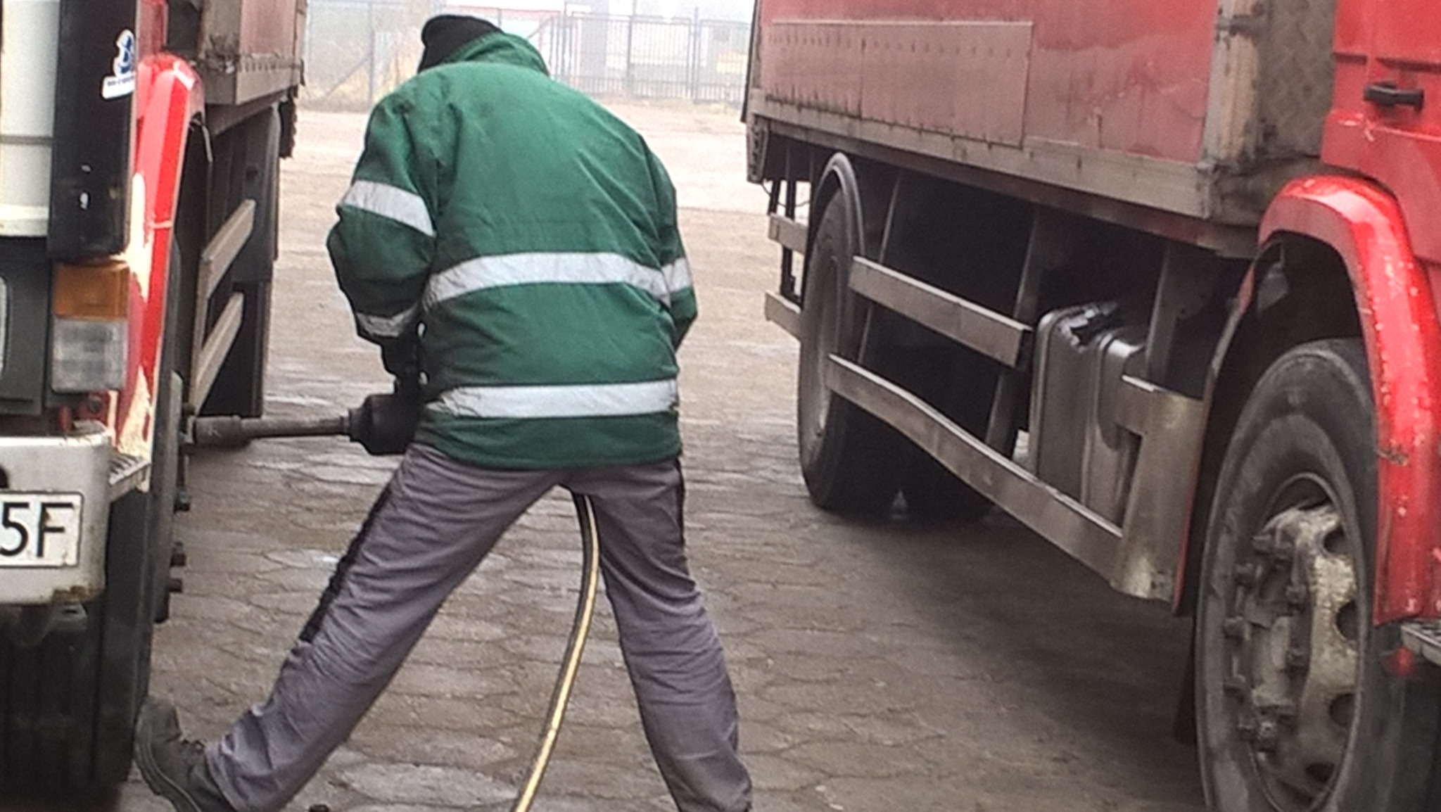 Mechanik w zielonej kurtce z białymi pasami, naprawia koło ciężarówki za pomocą klucza pneumatycznego, na tle zaparkowanych pojazdów i ogrodzenia.
