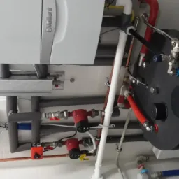 Nowoczesny, biały piec gazowy Vaillant z podgrzewaczem wody i instalacją CO w pomieszczeniu z białymi płytkami. Widoczne rury, pompy obiegowe i zawory.