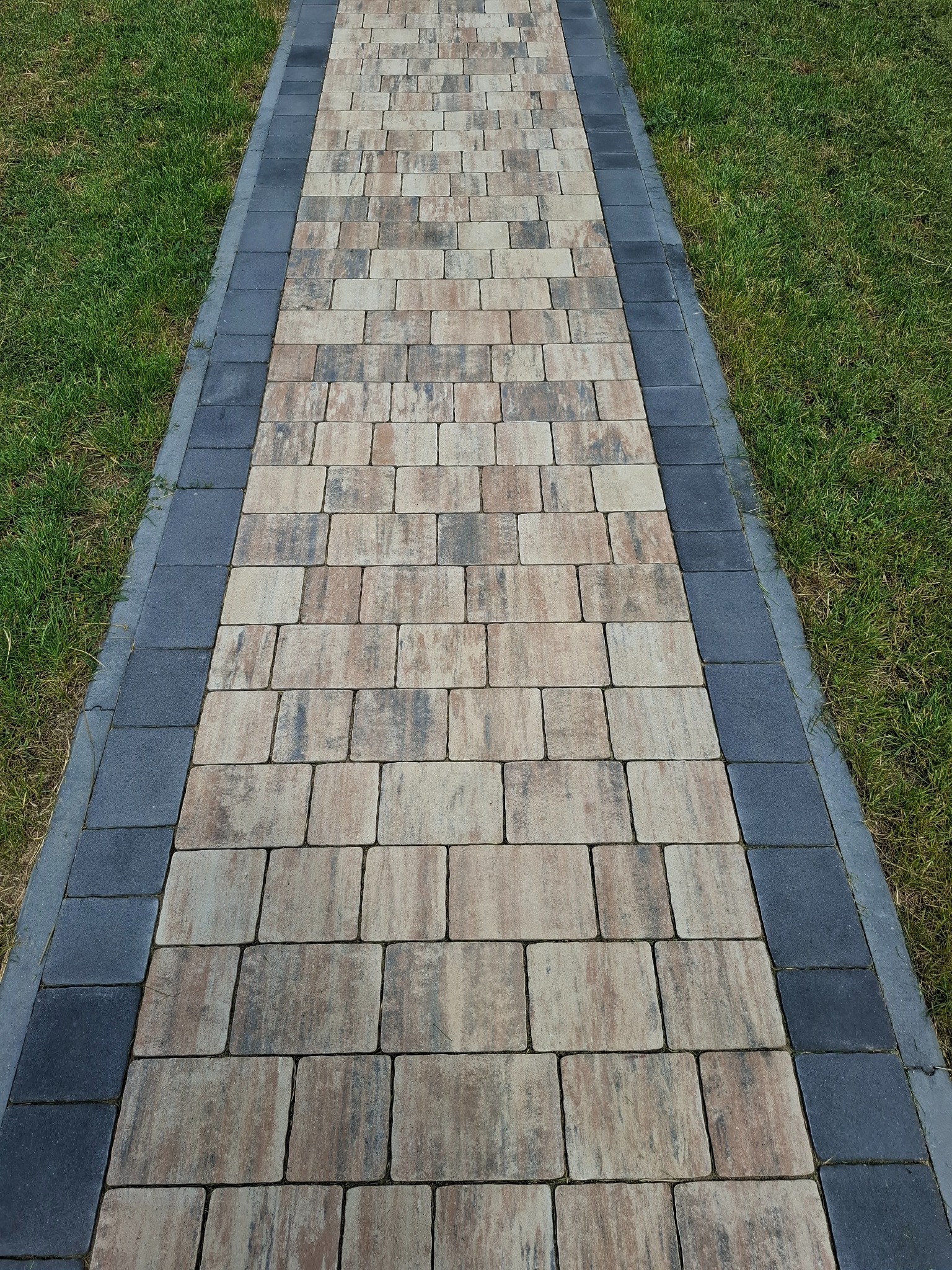 Ścieżka z kostki brukowej w odcieniach beżu i brązu, obramowana ciemnoszarą kostką, otoczona zielonym trawnikiem. Perspektywa z dołu, podkreślająca regularny wzór.