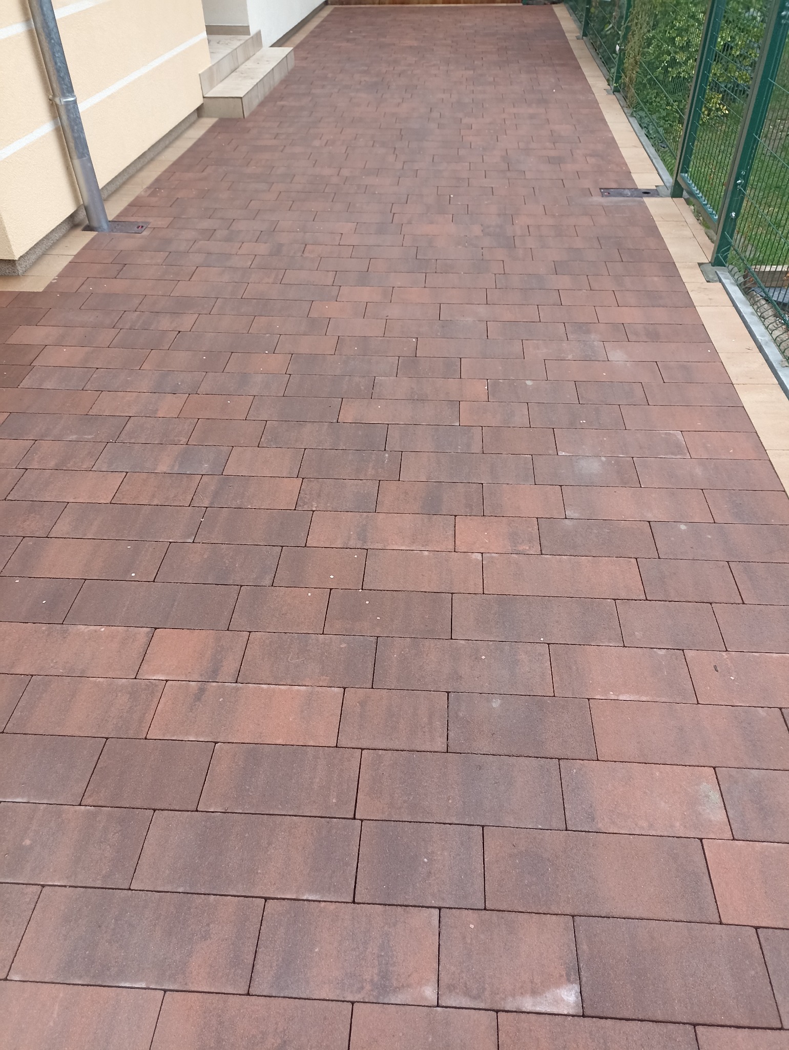 Ułożona, brązowa kostka brukowa na podjeździe, widok z góry. Metalowa rynna przy ścianie budynku. Ogrodzenie z siatki w tle. Regularny wzór ułożenia kostki.