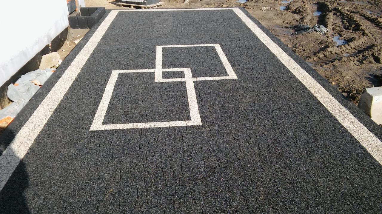 Czarna nawierzchnia z kostki z białymi obramowaniami i wzorem dwóch kwadratów, widoczny fragment budynku i błotniste podłoże w tle.