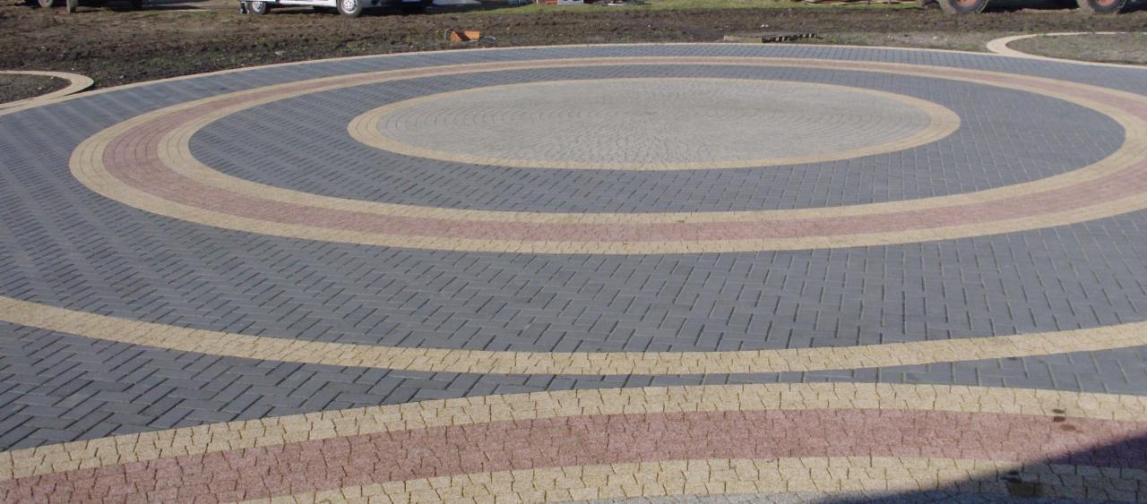 Okrągła powierzchnia z kostki brukowej ułożonej w koncentryczne wzory z szarej, beżowej i różowej kostki. W tle trawnik i samochód.