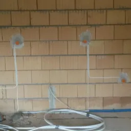 Instalacja elektryczna w trakcie montażu: przewody w ścianie z cegieł, puszki podtynkowe zalane gipsem, rura kanalizacyjna i zwój kabli na podłodze.