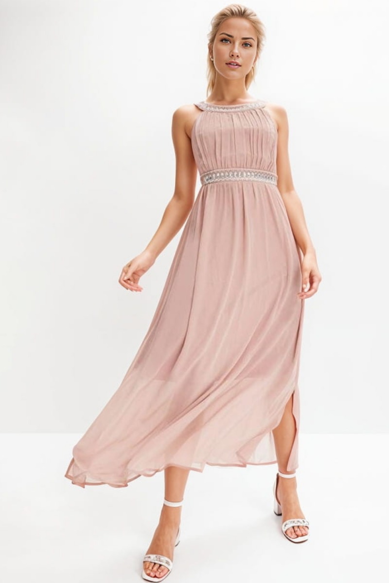 Kobieta pozuje w długiej, szyfonowej sukience w odcieniu brudnego różu z ozdobnym pasem i dekoltem, dopasowanej do białych sandałów na obcasie na białym tle.