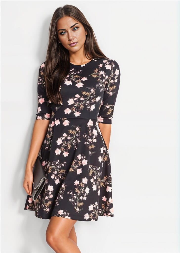 Kobieta prezentuje rozkloszowaną sukienkę mini z rękawem 3/4 i zabudowanym dekoltem, pokrytą wzorem w różowe kwiaty, trzymając czarną kopertówkę.