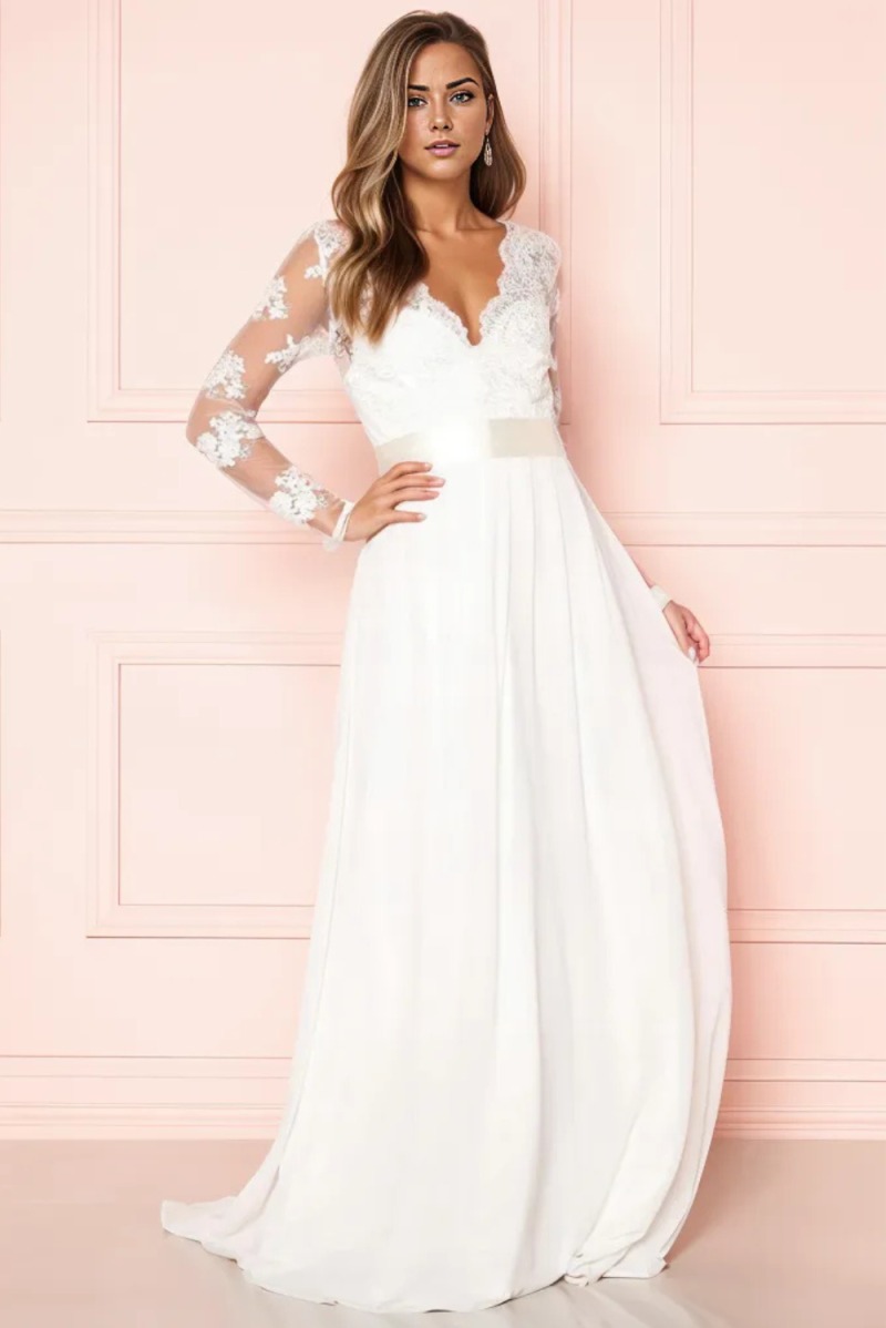 Kobieta w długiej, białej sukni ślubnej z koronkowymi rękawami i trenem, pozująca na tle ściany w pudrowym różu.
