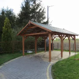 Drewniana konstrukcja z zadaszeniem, prawdopodobnie wiata garażowa, na posesji z kostką brukową i trawnikiem, otoczona zielenią.