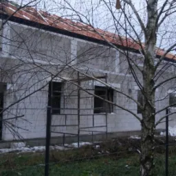 Surowy budynek w trakcie budowy z widocznym dachem z dachówki, otoczony drzewami i ogrodzeniem w zimowej scenerii.