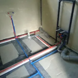 Rozprowadzenie instalacji wodnej i kanalizacyjnej w nowym budynku: rury czerwone i niebieskie ułożone na folii fundamentowej, szare rury kanalizacyjne, stelaż podtynkowy WC