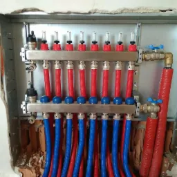 Rozdzielacz Purmo Premium z czerwonymi i niebieskimi rurami w skrzynce montażowej, widoczne zawory i przepływomierze, instalacja w trakcie montażu w surowym otoczeniu.