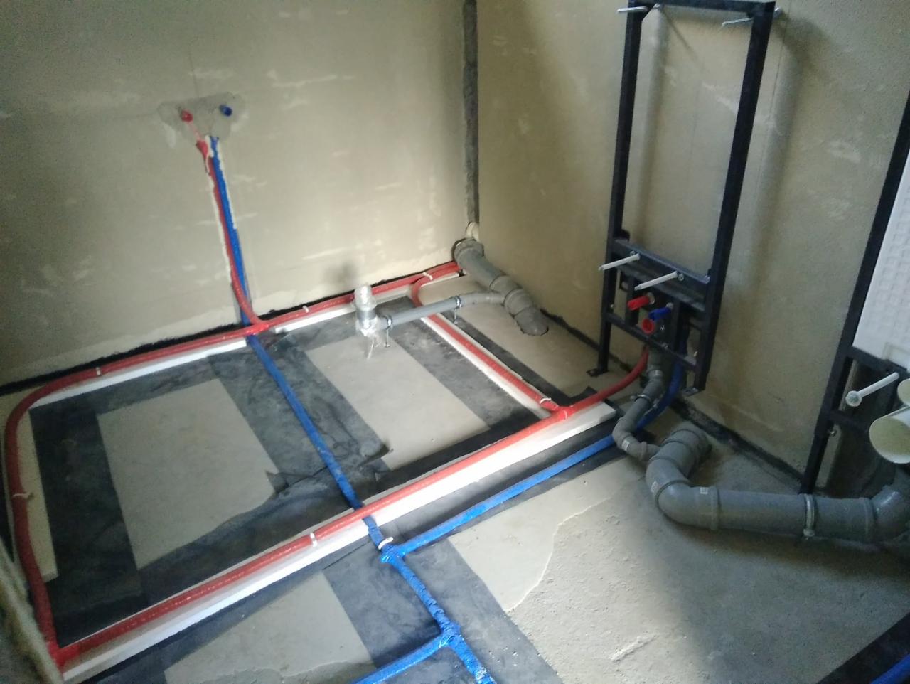 Rozprowadzenie instalacji wodnej i kanalizacyjnej w nowym budynku: rury czerwone i niebieskie ułożone na folii fundamentowej, szare rury kanalizacyjne, stelaż podtynkowy WC