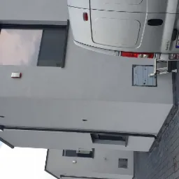 Szary budynek z zamontowanym czujnikiem alarmowym i skrzynką elektryczną na elewacji, częściowo zasłonięty przez srebrny samochód dostawczy z rozłożoną drabiną.