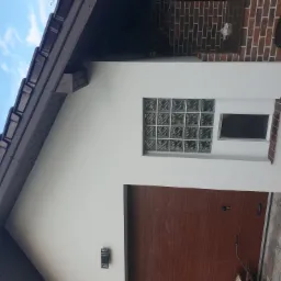 Ściana budynku z brązową bramą garażową, oknem z luksferów i lampą zewnętrzną, fragment dachu z brązową dachówką, na ścianie z cegły wisi grill.