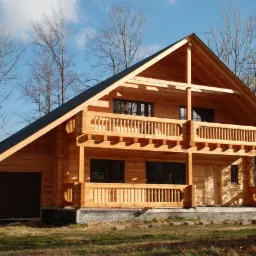 Dwukondygnacyjny dom z bali z balkonem i garażem, jasne drewno, widok z zewnątrz, błękitne niebo z chmurami w tle, drzewa bez liści i jedno drzewo z żółtymi liśćmi.