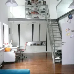 Nowoczesne wnętrze z antresolą, białe szafy z czarnymi wstawkami, metalowe schody i drewniana podłoga. Na antresoli szklana balustrada i półki z dekoracjami.