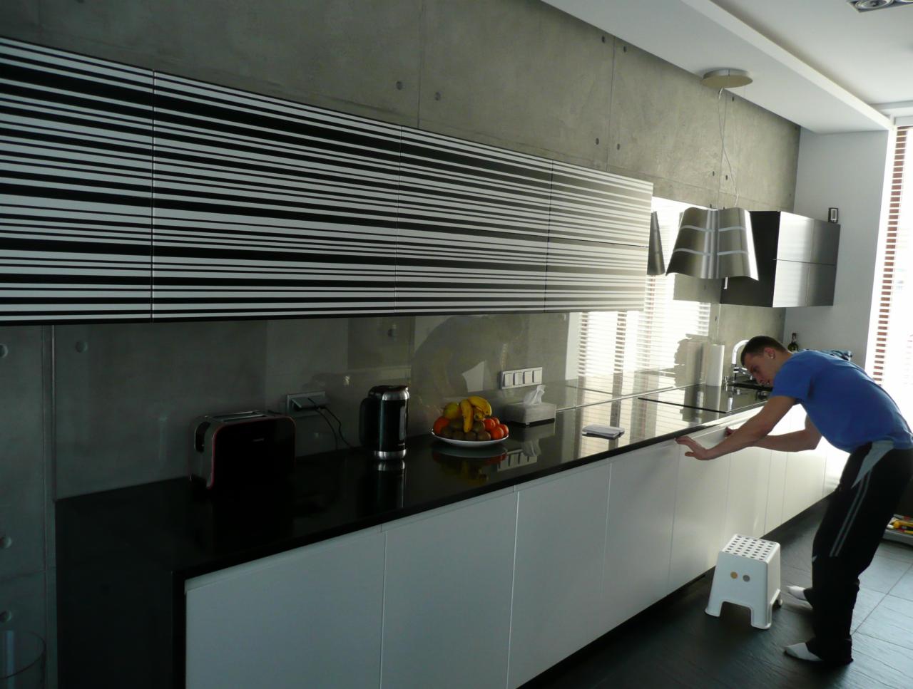 Nowoczesna kuchnia z czarnym blatem i szafkami w biało-czarne pasy, oświetlona lampą wiszącą. Mężczyzna w niebieskiej koszulce przy blacie. Minimalistyczny design.