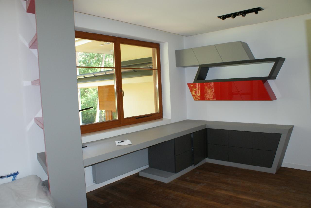 Nowoczesne meble na wymiar w szarościach i czerwieni, zintegrowane z oknem. Minimalistyczne półki na ścianie i drewniana podłoga dodają charakteru wnętrzu.