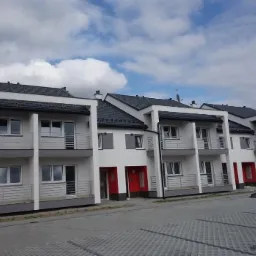 Rząd nowo wybudowanych, szeregowych domów dwukondygnacyjnych z balkonami, pomalowanych na biało z szarą podmurówką i ciemnymi dachami, czerwone drzwi wejściowe, kostka brukowa na podjeździe...