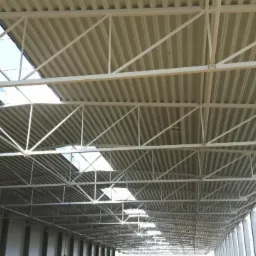 Wnętrze hali przemysłowej z widoczną konstrukcją dachu z blachy trapezowej i stalową kratownicą, oświetloną przez świetliki dachowe.