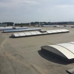 Rozległy, płaski dach przemysłowy z szarym pokryciem, wyposażony w szereg świetlików tunelowych z białymi ramami i przezroczystymi panelami, wentylatory dachowe, system odgromowy oraz widoczne...