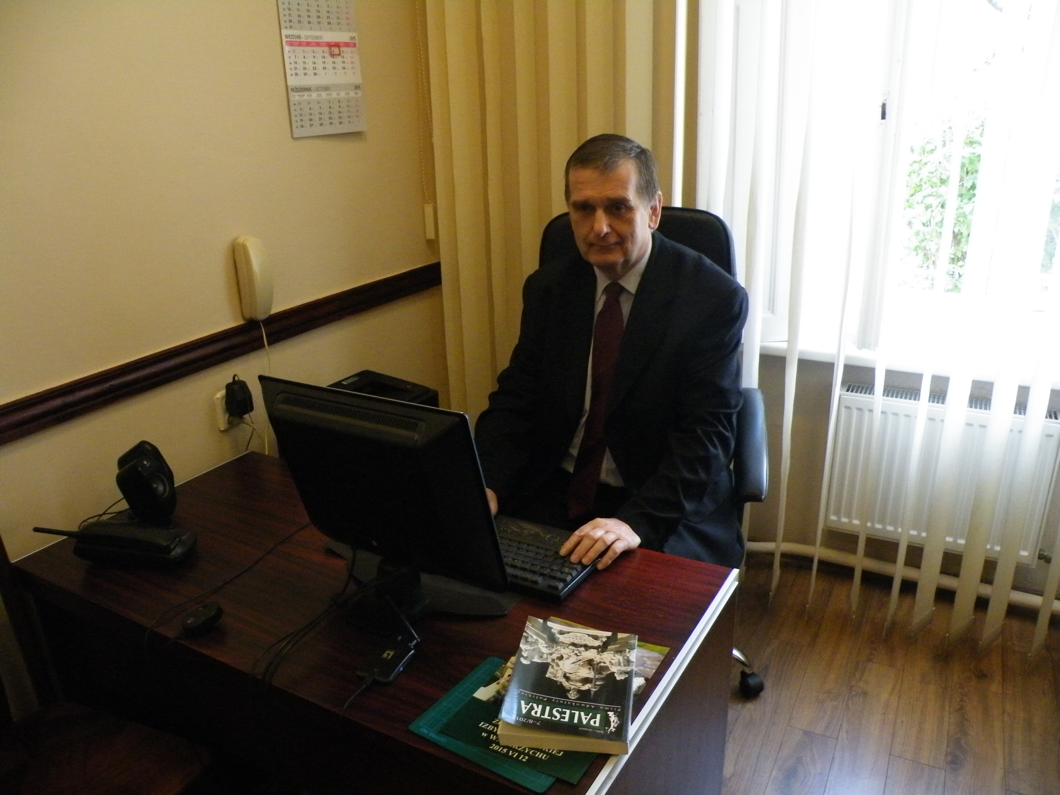 Adwokat w biurze, siedzi przy biurku z komputerem i książką 'Palestra', kalendarz na ścianie. Ubrany w garnitur i krawat. Naturalne światło z okna.