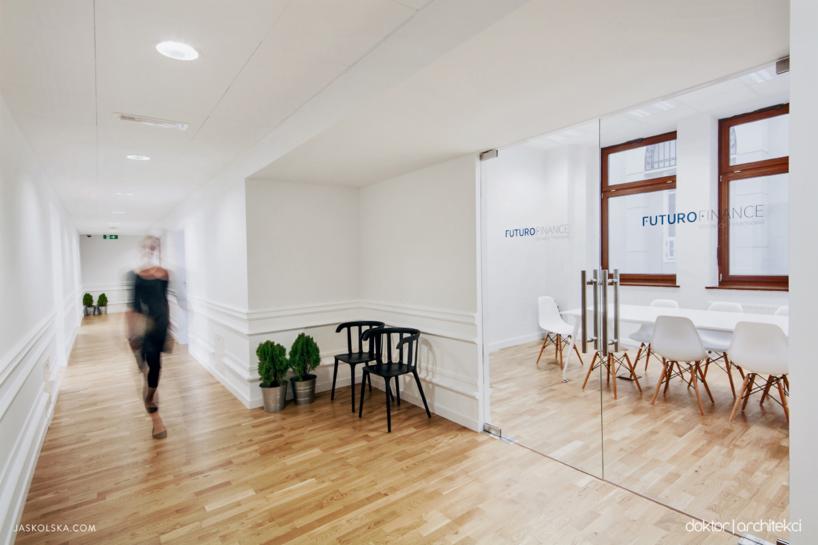 Białe wnętrze biura z drewnianą podłogą, przeszkloną salą konferencyjną z napisem 'Futuro Finance' i rozmytą sylwetką kobiety w przejściu.
