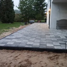 Ułożona szara kostka brukowa na posesji z widocznym fragmentem budynku, ławką i drzewami w tle, obrzeża kostki wykończone czarnym elementem, widoczny piasek przed kostką.