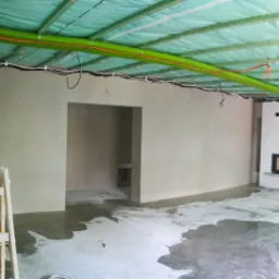 Wnętrze w trakcie prac wykończeniowych; świeżo otynkowane ściany, mokra posadzka, widoczna drabina malarska i instalacje pod sufitem.