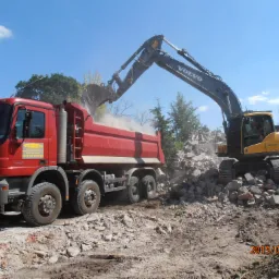 Żółta koparka Volvo załadowuje gruz na czerwoną ciężarówkę Mercedes Actros na placu rozbiórki pod błękitnym niebem. Data w prawym dolnym rogu: 2015/07/03.