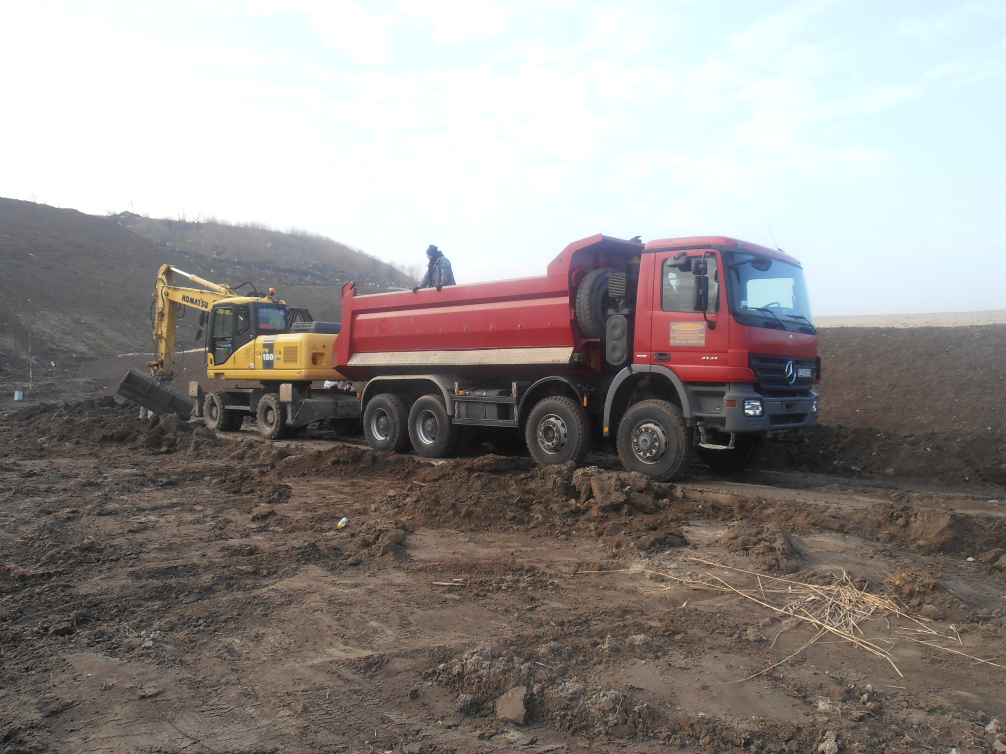 Czerwona ciężarówka Mercedes i żółta koparka Komatsu na placu budowy. Na skrzyni ciężarówki stoi osoba w niebieskiej kurtce. W tle widać usypany wał ziemi.