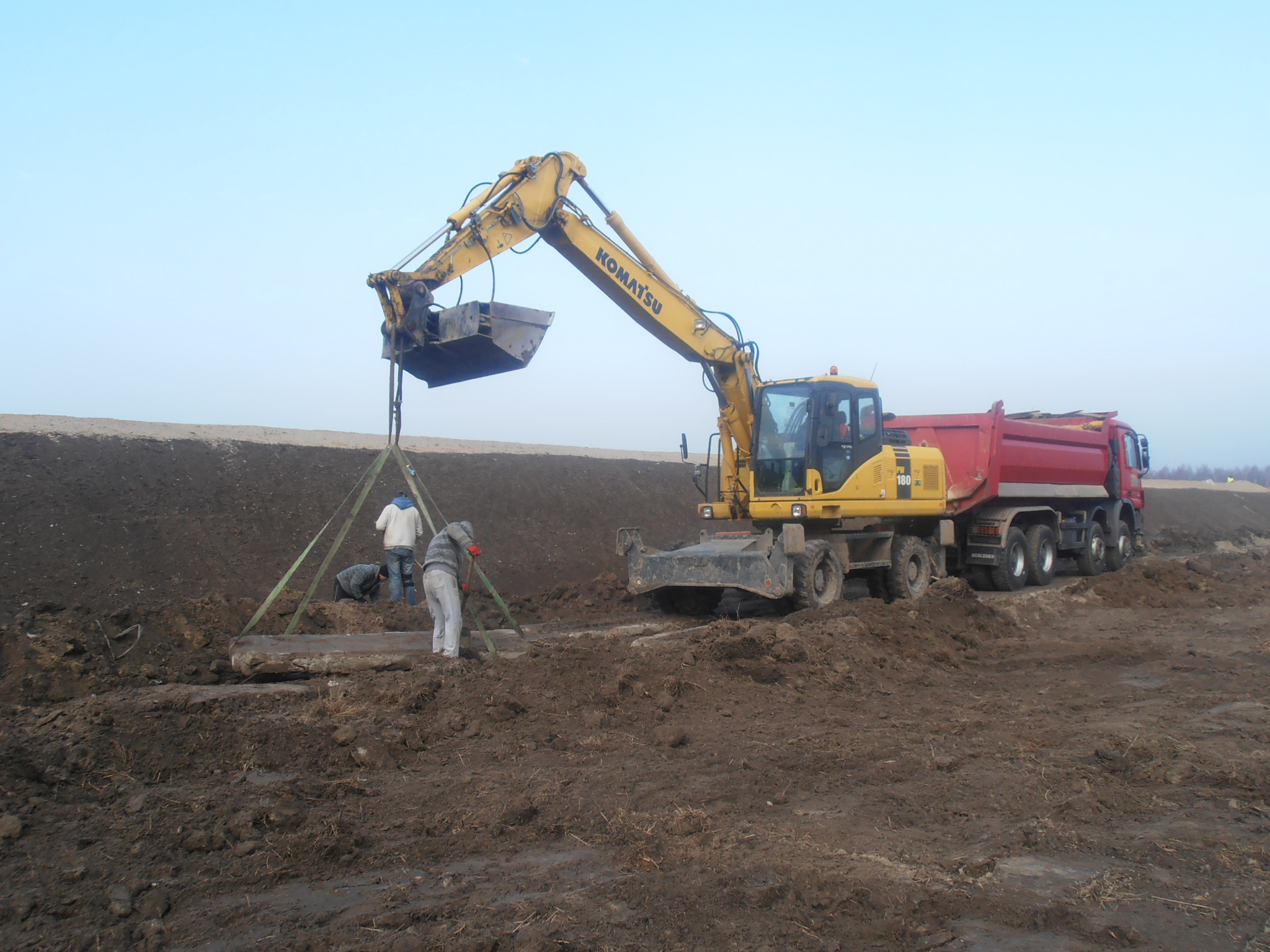 Żółta koparka Komatsu podnosi betonowy element na placu budowy. W tle czerwona ciężarówka i dwóch robotników. Niwelacja terenu.