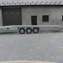 Długa, srebrna laweta z trzema osiami, zaparkowana na brukowanym placu przed budynkiem z szarymi drzwiami garażowymi i oknami.